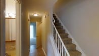 This multi-level space is thoughtfully laid out across three floors, offering a functional and well-organized floor plan. The layout, visible in the overhead model, reveals a series of interconnected rooms and open areas that provide flexibility in use and flow.

A central hallway serves as the main artery of the space, linking the various rooms with a clean, straightforward path. The hallway is finished with tile flooring and is illuminated by ceiling-mounted lighting, creating a welcoming corridor. At the end of the hallway, a door leads to a room that benefits from natural light filtering through a window, adding brightness to the interior.

A carpeted staircase with classic white balustrades and wooden newel posts connects the floors, offering a traditional aesthetic. The stair treads are finished in a neutral, earthy tone that complements the surrounding walls, which are painted in a soft, warm beige throughout.

The main open-plan area is spacious, featuring neutral flooring and ample room for a variety of configurations. Partition walls define separate functional zones while maintaining an open feel. Additional rooms branch off from the central areas, providing dedicated spaces for a range of uses. Overall, the space presents a practical, neutral canvas well-suited for customization.
