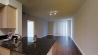This single-floor space showcases an open-concept design that seamlessly connects the kitchen and living areas. The kitchen features dark granite countertops with a polished finish, providing a sleek and functional workspace. Rich, dark hardwood flooring extends throughout the space, adding warmth and a sense of cohesion to the layout.

The living area is bathed in natural light, courtesy of large floor-to-ceiling windows dressed with vertical blinds, which allow for flexible light control. A track lighting fixture overhead provides additional illumination, ensuring the space remains well-lit at all times. The neutral gray walls complement both the dark flooring and countertops, creating a balanced and modern aesthetic.

A doorway visible from the main living area leads to an additional room, suggesting functional separation of spaces within the unit. The textured ceiling adds subtle visual interest overhead. Overall, the space presents a clean, contemporary atmosphere with a practical open layout well-suited for everyday living.