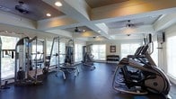 This fitness center is thoughtfully designed to provide a comprehensive workout environment. The space features an elegant coffered ceiling with recessed lighting and ceiling fans, lending both visual interest and comfortable air circulation during workouts. Large windows line the walls, flooding the interior with natural light and offering views of the surrounding greenery, creating an inviting and energizing atmosphere.

The rubber-tiled flooring provides durability and safety underfoot, ideal for the range of exercise equipment spread throughout the space. Strength training is well-supported with multiple cable and weight machines, including a multi-functional Smith machine and a cable crossover unit, as well as a dedicated free weight area with a full rack of dumbbells. On the opposite side, a row of cardio machines — including treadmills and other aerobic equipment — lines the perimeter, making efficient use of the available floor space.

Mirrors are positioned along the walls to assist with form and to visually expand the room. A framed piece of artwork adds a subtle decorative touch to the otherwise functional setting. Overall, the space strikes a balance between practicality and comfort, offering a well-rounded facility suitable for a variety of fitness routines.