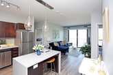 This contemporary single-floor residence showcases a thoughtfully designed layout with modern finishes throughout. The kitchen is a standout feature, equipped with rich dark espresso cabinetry, sleek bar-pull hardware, granite-style countertops, a subway tile backsplash, and a full suite of stainless steel appliances including a range, over-the-range microwave, dishwasher, and refrigerator. Wood-look flooring ties the kitchen together with a clean, polished aesthetic.

The living area is bright and inviting, featuring large sliding glass doors that open to a balcony with tree-lined outdoor views, filling the space with natural light. The room comfortably accommodates generous seating arrangements and is accented by wood-style flooring.

The bedrooms offer plush carpeted floors, ceiling fans, and large windows that frame lush outdoor scenery. Each bedroom provides a peaceful, comfortable retreat with ample natural light.

The bathroom is finished with wood-look flooring, a dark vanity with granite-style countertops, a large mirror with overhead lighting, and a full tub-shower combination with white tile surround. A hallway connects the bathroom directly to the bedroom, enhancing the flow of the space. Overall, the residence blends contemporary design with functional living.