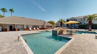 Located at 10480 Auto Mall Pkwy in D'Iberville, Mississippi, this outdoor amenity area offers a resort-style pool experience designed for relaxation and social gathering. The centerpiece is a generously sized swimming pool with crystal-clear water, ranging from shallow wading areas to deeper sections, complemented by a decorative tiled water feature that adds a dynamic focal point to the space.

The expansive pool deck is finished with attractive paver stonework and is furnished with lounge chairs positioned to take full advantage of the abundant natural sunlight. Towering palm trees and mature landscaping surround the area, enhancing the tropical ambiance and providing natural shade. A dedicated seating area features outdoor dining tables shaded by vibrant yellow umbrellas, creating an inviting space for casual gatherings.

A built-in outdoor grill station, constructed with stacked stone and equipped with a full-size stainless steel grill, is nestled within lush greenery, offering a convenient outdoor cooking area. The pool area is enclosed by decorative metal fencing for security and privacy, with gated access points. The adjacent single-story clubhouse building features large windows, a covered porch, and brick accents, providing a functional backdrop to this well-appointed outdoor retreat.