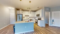 This single-floor residence offers a thoughtfully designed open-concept layout highlighted by light wood-tone luxury vinyl plank flooring that flows throughout the main living areas. The kitchen serves as the centerpiece of the space, showcasing crisp white shaker-style cabinetry, a classic white subway tile backsplash, and light stone-finish countertops. A large center island with a powder-blue base provides additional workspace and casual seating. Stainless steel appliances, including a French door refrigerator, electric range, and microwave, complete the kitchen. Two striking geometric brass-framed pendant lights hang above the island, adding a contemporary design element. The open living and dining areas are bathed in natural light from multiple windows and a glass-panel exterior door. Bedrooms feature plush carpeting, ceiling fans, and ample natural light, with at least one room offering a walk-in closet with built-in shelving. The bathroom includes a single-sink vanity with white cabinetry, stone-topped counters, a framed mirror, and a tub-shower combination. A dedicated laundry area houses full-sized washer and dryer units with overhead shelving for added storage convenience.