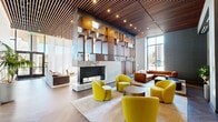 This upscale lounge in Bethesda, MD, offers a generously sized, single-floor gathering space defined by bold contemporary architecture and refined interior design. The centerpiece of the room is a dramatic marble fireplace framed by a towering sculptural wall of interlocking wood and metal geometric boxes, creating a striking focal point that draws the eye upward. Slatted warm wood ceilings run throughout, complemented by recessed lighting and cascading globe pendants that provide an inviting ambiance at any hour. Floor-to-ceiling windows and sheer white drapery wrap the perimeter, flooding the interior with abundant natural light while offering views of the surrounding Bethesda streetscape. The herringbone-patterned hardwood flooring adds texture and warmth underfoot. Distinct seating areas are thoughtfully arranged throughout, including clusters of vibrant chartreuse accent chairs, plush tufted sofas, and gold-accented side tables. A billiards table anchors a secondary social zone bathed in daylight. A media area with a wall-mounted display and burl wood console offers a more intimate retreat. The space also extends to a covered outdoor terrace with seating, planters, and open views of the neighborhood, seamlessly blending indoor comfort with outdoor accessibility.