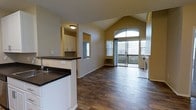 This single-floor space showcases an open-concept layout that seamlessly connects the kitchen and living areas. The most striking architectural feature is the vaulted ceiling, which rises to a dramatic peak and creates a strong sense of height and openness throughout the main living area. Dark hardwood-style flooring runs continuously across the space, providing a warm, grounding contrast to the neutral beige walls and white trim.

The kitchen is semi-open, separated from the living area by a raised breakfast bar with dark countertops and a white base. White cabinetry lines the kitchen walls, offering ample storage, while a pass-through window connects the kitchen to the rest of the living space, enhancing the open feel.

The living area is bathed in natural light, courtesy of a large arched window and a sliding glass door that leads to an outdoor balcony or patio. The arched window, positioned at the peak of the vaulted ceiling, serves as a focal point and frames outdoor views effectively. A wall-mounted HVAC unit is also present along the interior wall. Overall, the space feels bright, airy, and well-designed for both comfort and functionality.