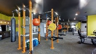 This fitness facility is designed to support a comprehensive strength and conditioning experience. The training floor is anchored by a series of bold orange power racks from Dynamic, each fully loaded with barbells, weight plates, and resistance bands, providing ample stations for squats, bench press, and a wide range of barbell movements. Stability balls in multiple sizes are stored directly on the racks for convenient access. The flooring features a durable, textured carpet-style surface well suited for heavy training activity. Large wall-mounted mirrors line the space, enhancing the sense of openness and allowing users to monitor their form throughout workouts. Overhead, a dark ceiling fitted with track lighting and ceiling fans ensures the space remains well-lit and ventilated during intense sessions. An extensive dumbbell rack is visible toward the rear of the facility, offering a broad selection of free weights to accommodate all fitness levels. Accent walls in yellow-green add a vibrant, energizing atmosphere to the environment. The layout is thoughtfully organized to maximize floor space and equipment accessibility, making it a functional and motivating environment for serious training.