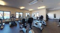 This fitness center offers a comprehensive workout environment designed to accommodate a variety of exercise routines. The space is laid out across a single floor and is divided into distinct functional zones, each catering to different training needs.

The main cardio area is generously sized and houses an extensive lineup of equipment, including upright and recumbent stationary bikes, treadmills, elliptical trainers, and stair climbers—many from the True Fitness brand. Multiple windows line the walls, allowing natural light to fill the room and providing outdoor views that enhance the overall atmosphere. Flush-mounted ceiling fixtures provide consistent overhead lighting throughout.

A dedicated strength training section features a multi-station cable machine, a leg press unit, and additional selectorized equipment, giving users options for full-body resistance training. A separate free weights area includes a fully stocked three-tier dumbbell rack, a workout bench, and a wall-mounted mirror for form checks. Wall-mounted flat-screen televisions are positioned in multiple areas of the facility for entertainment during workouts. Dark rubberized flooring throughout the space provides durability and traction underfoot.