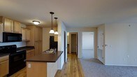 This single-floor residence offers a practical and well-organized layout designed for comfortable everyday living. The kitchen serves as a central hub of the space, featuring light wood cabinetry, dark countertops, and a stainless steel sink set into a peninsula island. Two pendant lights hang above the island, providing focused task lighting while adding a stylish touch to the kitchen's overall aesthetic. A black refrigerator anchors the appliance lineup, complementing the dark countertop tones. The kitchen transitions seamlessly into the adjoining living areas, where the flooring shifts from warm hardwood to plush carpet, delineating the spaces naturally. A hallway leads to additional rooms, including what appears to be a bedroom with a walk-in closet and a bathroom, offering a thoughtful and functional layout. Neutral wall tones throughout the residence lend a clean, move-in-ready feel, while standard ceiling heights maintain a comfortable sense of space. The overall ambiance is practical and welcoming, with a cohesive design palette of warm wood tones, neutral walls, and dark accents that tie the space together cohesively across all rooms.