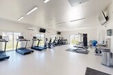 Located at 2659 Ruff Rd in Jackson, Michigan, this single-story fitness center offers a well-equipped and thoughtfully designed space for exercise and wellness. The facility comprises a welcoming entrance area and a generously sized main exercise room, all presented in a clean, bright aesthetic with crisp white walls and light-toned flooring throughout.

The entrance area provides a functional transition into the facility, featuring seating along the wall, a water fountain station, and a coat rack — practical amenities that set a comfortable tone upon arrival. A mounted flat-screen television and mirror add to the welcoming atmosphere.

The main exercise room is open and airy, benefiting from abundant natural light streaming through multiple large sliding glass doors and windows that line the walls, offering views of the surrounding greenery. High ceilings and recessed linear lighting further enhance the sense of spaciousness. The room is well-ventilated with wall-mounted climate control units.

Cardio equipment is prominently featured, including multiple treadmills, elliptical trainers, and stationary bikes arranged in organized rows. A multi-station cable strength machine with a flat bench provides options for resistance training, while a dedicated free weights area with a dumbbell rack and floor mats rounds out the strength training offerings. Wall-mounted televisions are positioned throughout for entertainment during workouts.