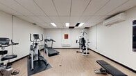 Located at 1300-1310 McWatters Rd in Ottawa, Ontario, this dedicated fitness room provides a well-equipped workout space within a compact, efficiently utilized footprint. The room features light wood-style flooring throughout, contributing to a clean and functional aesthetic. A suspended drop-tile ceiling with recessed panel lighting ensures consistent, even illumination across the entire space.

The fitness room is outfitted with a comprehensive selection of exercise equipment designed to support both cardiovascular and strength training. Cardio offerings include a treadmill, an elliptical trainer, and a recumbent stationary bike, all positioned to allow comfortable use. For strength training, the room includes a multi-function cable and weight machine, a flat weight bench, and a dedicated dumbbell rack stocked with a full range of free weights. Anti-fatigue rubber mats are placed beneath select equipment for safety and comfort.

The walls are finished in a neutral white tone, lending the space an open and airy feel. A wall-mounted split air conditioning unit ensures climate control year-round. Motivational fitness artwork is displayed on one wall, adding character to the room. A small shelving unit provides convenient storage for accessories. The space is accessed through a secure keyed entry door, reinforcing its role as a private, amenity-grade facility.