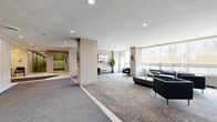 Located at 200 Rideau Terrace in Ottawa's desirable Rockcliffe Park neighbourhood, this elegantly designed residential lobby offers a welcoming and well-appointed entry experience. The generously sized single-floor space is finished with a combination of patterned carpet and clean tile flooring, guiding residents and visitors through distinct functional zones. Upon entry, an enclosed vestibule with automatic glass doors and polished stone-clad walls provides a refined first impression, with seating available for guests. The main lobby opens into a broad, open lounge area bathed in natural light from large windows fitted with roller shades. This seating area features contemporary dark leather furniture, a sofa, and armchairs arranged around a low coffee table, creating a comfortable and sophisticated gathering space. A sleek wall-mounted electric fireplace set against large-format stone tile serves as a focal point in an adjoining alcove, accompanied by additional lounge seating. Curated artwork and a tall indoor plant add warmth and character throughout. Recessed ceiling lighting illuminates the entire space evenly. Practical amenities include a dedicated mailroom with individual resident mailboxes, elevator access, and clearly marked building corridors leading further into the property. The overall ambiance is modern, calm, and professionally maintained.