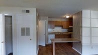 This single-floor space showcases a kitchen that serves as a central feature of the layout. The kitchen is equipped with warm-toned wood cabinetry spanning both upper and lower sections, providing ample storage. A tiled backsplash lines the countertops, which include an integrated sink positioned beneath the upper cabinets. White appliances, including a dishwasher and range, contribute a clean, classic aesthetic. A peninsula counter extends into the living area, creating a natural divide between spaces while offering additional workspace or casual seating. The flooring throughout is a dark hardwood-style material, adding warmth and visual continuity across the space. Walls feature a neutral, textured wallcovering that complements the overall color palette. A textured ceiling with flush-mount lighting provides functional illumination. HVAC vents are visibly integrated into the walls, ensuring climate control throughout. A white interior door near the entrance leads to an additional room or closet, contributing to the unit's practical layout. Overall, the space presents a straightforward, functional design well-suited for everyday living.
