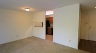 This single-floor space offers a practical and straightforward layout designed for comfortable everyday living. The living area is the central feature of the unit, finished with wall-to-wall carpeting that adds warmth underfoot. A pass-through opening connects the living area to the kitchen, allowing for a sense of openness between the two spaces. The kitchen is equipped with warm-toned wood cabinetry, stone-look countertops, and black appliances including a refrigerator and range, all set against a tile backsplash. The kitchen flooring transitions to a hard surface, providing a durable and easy-to-clean workspace. A hallway connects the main living areas to additional rooms, which appear to include a bathroom fitted with standard fixtures. The walls throughout the unit are finished in a clean, neutral white, contributing to a bright and open atmosphere. Ceiling-mounted lighting fixtures provide consistent illumination across the living spaces. The overall layout is efficient and well-defined, with clear separation between the kitchen and living zones while maintaining a cohesive flow throughout the single-level floor plan.