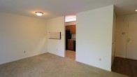 This single-floor space offers a practical and straightforward layout designed for comfortable everyday living. The living area is the central feature of the unit, finished with wall-to-wall carpeting that adds warmth underfoot. A pass-through opening connects the living area to the kitchen, allowing for a sense of openness between the two spaces. The kitchen is equipped with warm-toned wood cabinetry, stone-look countertops, and black appliances including a refrigerator and range, all set against a tile backsplash. The kitchen flooring transitions to a hard surface, providing a durable and easy-to-clean workspace. A hallway connects the main living areas to additional rooms, which appear to include a bathroom fitted with standard fixtures. The walls throughout the unit are finished in a clean, neutral white, contributing to a bright and open atmosphere. Ceiling-mounted lighting fixtures provide consistent illumination across the living spaces. The overall layout is efficient and well-defined, with clear separation between the kitchen and living zones while maintaining a cohesive flow throughout the single-level floor plan.