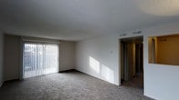 This single-floor space features a spacious living area finished with wall-to-wall carpet in a neutral tone, providing a comfortable and practical foundation throughout. A large sliding glass door fitted with vertical blinds dominates one wall, allowing generous natural light to filter into the room and casting distinct patterns across the interior. The blinds offer adjustable privacy and light control while providing access to an outdoor area beyond. The ceiling features a textured popcorn finish, and overhead HVAC vents are integrated into the ceiling for efficient climate control. The walls are painted in a clean, neutral white, contributing to a bright and open atmosphere. A doorway visible toward the rear of the living area leads to a hallway connecting to additional rooms beyond, suggesting a practical and straightforward layout. The overall ambiance is clean, understated, and well-lit, making it a versatile space suited to a variety of furnishing arrangements.