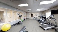 This fitness center offers a spacious, single-level layout designed to accommodate a wide range of exercise activities. The room features a drop-panel ceiling equipped with recessed fluorescent lighting panels that provide consistent, bright illumination throughout the space, ensuring excellent visibility during workouts. Natural light is supplemented through several windows along the walls, contributing to an open and inviting atmosphere.

The flooring consists of durable, light-toned carpet tiling, which provides cushioning underfoot and helps define the workout area. Along one side of the room, a row of cardio machines — including treadmills, ellipticals, and stationary bikes — are neatly arranged, several facing the windows. A wall-mounted television is positioned nearby for entertainment during cardio sessions.

The strength training area includes a multi-station cable machine, a dumbbell rack with a selection of free weights, and a dedicated stretching bench. Exercise accessories such as stability balls and medicine balls are also available. Mirrors are mounted along portions of the walls, enhancing the sense of depth and allowing users to monitor their form. The overall ambiance is clean, organized, and functional, reflecting a professionally maintained fitness environment.