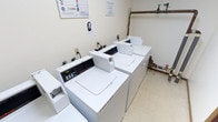 This shared laundry facility is a compact, single-floor utility space designed to serve the practical needs of its users. The room features multiple commercial-grade top-loading washing machines, including Speed Queen models, alongside matching dryers arranged in a row along the walls, providing ample capacity for laundry needs. The space is finished with light-colored walls and durable tile flooring, creating a clean and functional environment. Overhead fluorescent lighting ensures the room is well-lit throughout.

Exposed copper and PVC plumbing lines run along the walls, connecting to the machines and a water heater housed within the room, reflecting the facility's straightforward, utilitarian design. A cork bulletin board is mounted on the wall, displaying informational notices and laundry safety guidelines for users. Ventilation is provided via a ceiling vent, supporting air circulation within the enclosed space. A doorway with wood trim provides access to an adjacent carpeted hallway. The overall ambiance is practical and no-frills, prioritizing functionality and ease of use for its occupants.