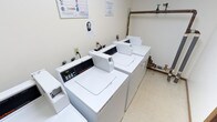 This shared laundry facility is a compact, single-floor utility space designed to serve the practical needs of its users. The room features multiple commercial-grade top-loading washing machines, including Speed Queen models, alongside matching dryers arranged in a row along the walls, providing ample capacity for laundry needs. The space is finished with light-colored walls and durable tile flooring, creating a clean and functional environment. Overhead fluorescent lighting ensures the room is well-lit throughout.

Exposed copper and PVC plumbing lines run along the walls, connecting to the machines and a water heater housed within the room, reflecting the facility's straightforward, utilitarian design. A cork bulletin board is mounted on the wall, displaying informational notices and laundry safety guidelines for users. Ventilation is provided via a ceiling vent, supporting air circulation within the enclosed space. A doorway with wood trim provides access to an adjacent carpeted hallway. The overall ambiance is practical and no-frills, prioritizing functionality and ease of use for its occupants.