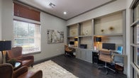 This single-floor office space combines functionality and refined design, offering a polished environment well-suited for professional use. The room is anchored by an expansive built-in workstation wall finished in a muted gray tone, featuring multiple desk surfaces, under-counter cabinetry, and open display shelving with accent lighting—providing ample workspace for two individuals simultaneously. Dark hardwood floors run throughout, adding warmth and contrast against the light gray walls and crisp white ceilings accented with crown molding. Recessed lighting ensures consistent illumination across the space, complemented by task lamps at each workstation. A dedicated seating area near the window features two rich brown leather wingback chairs arranged around a brass accent table, creating an inviting space for meetings or casual conversation. Natural light enters through a large window fitted with wood-tone blinds, offering a connection to the outdoors while maintaining privacy. Abstract artwork adorns the walls, adding a contemporary aesthetic touch throughout. A glass-paneled interior door provides visual openness while maintaining a degree of separation, enhancing the professional character of the space. An ornamental area rug anchors the seating zone, tying together the room's neutral and earthy color palette.