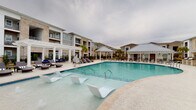 This outdoor pool area in Youngsville, Louisiana, is designed to deliver a resort-style experience within a well-appointed residential community. The centerpiece is a generously sized swimming pool with crystal-clear water, featuring in-pool lounge chairs that allow residents to relax while partially submerged. The expansive surrounding deck is finished in smooth, light-toned concrete, offering ample space for sunbathing and socializing, complemented by a collection of cushioned chaise lounges and wicker outdoor seating with navy blue cushions.

A covered pavilion with a classic white pergola anchors one end of the pool area, providing shaded retreat from the Louisiana sun. An additional covered structure built with warm brick and white trim serves as a poolside clubhouse, featuring large glass French doors, an outdoor mounted flat-screen television, and a ping pong table for recreational use. Pool rules are clearly posted, and the space is enclosed by a black iron fence for safety and privacy.

Mature palm trees and manicured landscaping add a tropical ambiance throughout the courtyard. The multi-story residential buildings surrounding the pool are constructed with a blend of brick and painted siding, their balconies overlooking the amenity area. The overall atmosphere is vibrant yet relaxed, making this pool deck a standout outdoor amenity.