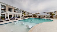 This outdoor pool area in Youngsville, Louisiana, is designed to deliver a resort-style experience within a well-appointed residential community. The centerpiece is a generously sized swimming pool with crystal-clear water, featuring in-pool lounge chairs that allow residents to relax while partially submerged. The expansive surrounding deck is finished in smooth, light-toned concrete, offering ample space for sunbathing and socializing, complemented by a collection of cushioned chaise lounges and wicker outdoor seating with navy blue cushions.

A covered pavilion with a classic white pergola anchors one end of the pool area, providing shaded retreat from the Louisiana sun. An additional covered structure built with warm brick and white trim serves as a poolside clubhouse, featuring large glass French doors, an outdoor mounted flat-screen television, and a ping pong table for recreational use. Pool rules are clearly posted, and the space is enclosed by a black iron fence for safety and privacy.

Mature palm trees and manicured landscaping add a tropical ambiance throughout the courtyard. The multi-story residential buildings surrounding the pool are constructed with a blend of brick and painted siding, their balconies overlooking the amenity area. The overall atmosphere is vibrant yet relaxed, making this pool deck a standout outdoor amenity.