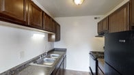 This single-story space offers a clean, straightforward layout designed for comfortable everyday living. The living area is spacious and open, finished with neutral-toned wall-to-wall carpeting that adds warmth underfoot. Two windows along the exterior wall allow natural light to fill the room, creating a bright and airy atmosphere. A ceiling fan is centrally positioned, providing both lighting and ventilation. The neutral wall color throughout gives the space a calm, versatile backdrop suitable for a variety of furnishing styles.

The kitchen is efficiently laid out with a galley-style design that maximizes functionality. Rich, dark wood cabinetry lines both the upper and lower sections of the walls, providing ample storage space. Granite-style countertops with a double stainless steel sink offer practical workspace, while the electric range with an overhead ventilation hood accommodates everyday cooking needs. Light tile flooring contrasts neatly with the darker cabinetry. Overhead lighting keeps the kitchen well-illuminated. Overall, the space presents a functional and tidy environment with a cohesive neutral palette throughout.