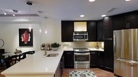 This single-story space showcases a well-appointed modern kitchen designed with both style and functionality in mind. The kitchen is anchored by rich, dark espresso-toned cabinetry that extends from floor to ceiling, providing generous storage throughout. The cabinetry is complemented by crisp white countertops, creating a striking visual contrast that gives the space a clean, contemporary feel.

A peninsula with an integrated undermount sink serves as a central feature, offering additional workspace and casual seating potential. Pendant lighting with chrome fixtures hangs above the peninsula, adding warmth and a refined decorative touch alongside recessed ceiling lights that ensure the space is well illuminated throughout.

Stainless steel appliances, including a slide-in range with oven, an over-the-range microwave, and additional built-in units, reinforce the kitchen's modern aesthetic. Light-toned tile backsplash lines the walls between the upper and lower cabinets, adding subtle texture without disrupting the cohesive color palette. The hardwood flooring anchors the space warmly, tying together the dark cabinetry and bright surfaces for a balanced, sophisticated overall ambiance.