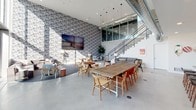 Located at 13448 Beach Ave in Los Angeles, CA 90066, this single-floor space delivers a thoughtfully designed environment that blends industrial aesthetics with modern comfort. The main common area commands attention with soaring double-height ceilings, exposed ductwork, and polished concrete floors that lend an authentic industrial character. A bold patterned feature wall anchors the lounge zone, where eclectic seating arrangements — including upholstered chairs and intimate café-style tables — invite both collaboration and casual gathering. A large communal wooden table with mixed seating serves as a central focal point, ideal for group work or social interaction. A wall-mounted multi-panel display adds a media-ready element to the space. An open staircase with glass paneling leads to a mezzanine level overlooking the main floor, adding architectural depth and visual interest. Pendant lighting with geometric cage fixtures illuminates the kitchen, which features warm wood cabinetry, dark stone countertops, a speckled tile backsplash, a professional-grade range, and a central island with bar seating — making it both functional and refined. Natural light filters throughout, enhancing the bright, airy atmosphere of this versatile Los Angeles property.