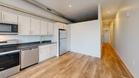 Located at 2239 Railroad St in Pittsburgh's dynamic Strip District, this one-bedroom, one-bathroom unit offers a thoughtfully designed single-floor layout with modern industrial character. Light-toned hardwood-style flooring runs throughout the space, complementing the clean white walls and creating a cohesive, contemporary aesthetic.

The open-concept kitchen is a standout feature, outfitted with white cabinetry, dark granite-style countertops, and a full suite of stainless steel appliances including a refrigerator, range, microwave, and dishwasher. Exposed silver ductwork along the ceiling adds an industrial loft feel that distinguishes the space.

The bedroom is bright and airy, benefiting from abundant natural light streaming through large sliding glass doors that open onto a private balcony. The balcony offers open views of the Pittsburgh skyline, providing a compelling outdoor extension of the living space.

A hallway connects the main living areas to the bathroom and a built-in closet with sliding doors, optimizing storage within the layout. The bathroom is finished with a dark granite vanity top, light cabinetry, and wood-look tile flooring, maintaining the consistent modern design language found throughout the unit. Recessed lighting throughout ensures well-illuminated interiors day and night.