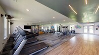 This fitness center in Baton Rouge, Louisiana offers a comprehensive, single-floor facility designed to support a wide range of workout routines. The main workout area features wood-look flooring and is lined with strength training equipment, including a dumbbell rack, weight benches, cable machines, and a multi-function rack — all reflected by a full wall of mirrors that amplifies the sense of space. Recessed lighting and linear LED fixtures illuminate the facility throughout.

A dedicated cardio section houses a row of treadmills positioned beneath large windows, allowing natural light and exterior views to create an open, energizing atmosphere. Wall-mounted fans provide additional airflow in this area. Nearby, stationary bikes, a rowing machine, and additional cardio equipment are arranged in an open zone alongside a flat-screen TV, medicine balls, and resistance training accessories.

A separate yoga and stretching room provides a quieter, more meditative environment, featuring a colorful yoga-themed wall mural, exercise mats, stability balls, and a BOSU trainer. The room's understated design encourages focus and mindfulness.

Vibrant "Exercise" and "Wellness" word art murals flank the main entrance double doors, which open to outdoor green space, reinforcing the facility's commitment to an active, health-forward lifestyle. The overall ambiance is modern, clean, and motivating.