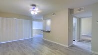 This single-story residence offers a thoughtfully designed open-concept layout that maximizes the sense of space and flow. The main living area features smooth wood-look flooring that extends throughout, complemented by neutral-toned walls and crisp white baseboards that give the interior a clean, contemporary feel. A ceiling fan with a light fixture provides both comfort and ambient lighting to the living space, while a large window fitted with horizontal blinds allows natural light to fill the room.

A partial wall with a breakfast bar counter serves as a stylish divider between the living area and the kitchen, creating an open connection while maintaining defined zones. The kitchen is equipped with a stainless steel refrigerator and a sink visible from the living space, offering practical functionality alongside an open feel. A textured popcorn ceiling adds a classic architectural detail overhead, consistent throughout the space.

Vertical blinds adorn a sliding door or large window along one wall, hinting at access to an outdoor area. The overall ambiance is bright, clean, and move-in ready, with a neutral color palette that provides a versatile backdrop suited to a variety of interior styles. The single-floor layout ensures easy accessibility and seamless navigation throughout the home.
