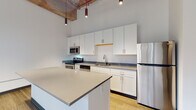Located at 366 E Broad St in the heart of Columbus, Ohio, this single-floor loft-style unit blends industrial character with contemporary finishes. Soaring ceilings anchored by exposed wooden beams set an airy, open tone throughout the space, while pendant lighting adds a modern, intentional design touch.

The kitchen is a standout feature, offering a large center island with light quartz countertops, white shaker cabinetry extending to the ceiling, stainless steel appliances including a range, microwave, dishwasher, and refrigerator, and light hardwood flooring that enhances the bright, clean aesthetic. Ample counter and storage space make the kitchen both functional and visually appealing.

The carpeted bedrooms are generously proportioned, with tall ceilings that amplify the sense of space. Large, grid-paned windows in the bedroom areas allow natural light to filter in and offer views of the surrounding urban streetscape. The rooms connect seamlessly through corridor hallways with dark-trimmed doorframes that complement the overall design palette.

The bathroom is finished with neutral large-format tile flooring, a white vanity with quartz countertop, a matte black faucet, and a tiled walk-in shower, delivering a clean and refined look. Throughout the unit, the interplay of raw architectural elements and polished modern finishes creates a distinctly urban loft atmosphere.
