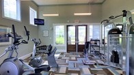 This fitness center at 19000 Oak Road West in Gulf Shores, Alabama, is a well-appointed single-story exercise facility designed to support a range of workout routines. The space features notably high ceilings that lend an open, airy feel, enhanced by large windows and glass double doors that invite abundant natural light while offering views of the surrounding greenery. The geometric-patterned flooring in neutral tones of blue, tan, and brown adds a modern aesthetic to the interior. Overhead fluorescent lighting ensures the space remains bright and functional at any hour. The facility is equipped with a variety of cardio machines, including a recumbent bike and additional exercise equipment, as well as a dedicated free weights area with a rack of dumbbells. Strength training equipment and resistance machines round out the offerings, making the space suitable for a full-body workout. A wall-mounted television provides entertainment during exercise sessions. Mirror panels along one wall enhance the sense of space and support proper form during workouts. The soft green wall color contributes to a calm, focused atmosphere throughout the facility.