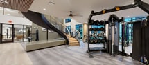 This upscale fitness center in Charlotte, NC spans 3,510 square feet across two floors — 1,720 sq ft on the ground level and 1,790 sq ft above — delivering a comprehensive workout environment with a refined, modern aesthetic. Light wood-plank flooring flows throughout both levels, complemented by warm wood-wrapped columns, recessed lighting, and wood-slat ceiling accents that add architectural warmth.

The upper floor features expansive floor-to-ceiling windows that flood the space with natural light and offer views of the surrounding community. This level houses a wide array of cardio and strength equipment, including True treadmills, stationary and spin bikes, rowing machines, functional trainers, cable machines, free weights, and a multi-functional power rack. Ceiling fans add comfort to the workout areas, and rubber flooring defines dedicated strength zones.

The lower level offers a versatile studio space with mirrored walls, a ballet barre, and a slatted wood ceiling — ideal for group classes or stretching. A reception-style entry area with built-in storage cubbies and curated artwork provides a welcoming transition. A private room with clean, minimalist finishes offers additional flexibility. The interconnected staircase with sleek metal cable railings unifies both levels into a cohesive, well-appointed fitness facility.