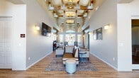 This generously sized single-story clubhouse facility offers an impressive array of communal spaces designed for both comfort and functionality. The entrance welcomes visitors with soaring double-height ceilings framed by an elegant coffered structure featuring warm wood beam accents and statement pendant lighting. Large arched transom windows flood the interior with abundant natural light, while hardwood-style flooring runs seamlessly throughout. The main lounge area is thoughtfully furnished with upholstered seating arrangements set atop decorative area rugs, creating an inviting atmosphere for gathering. Wall-mounted displays and curated artwork add a polished, refined character to the space.

The combined dining and living room area provides a casual setting complete with a coffee bar station, high-top tables, and additional seating, ideal for residents to relax or socialize. A dedicated office space offers a quiet, focused environment equipped with a workstation and complemented by soothing sage-toned walls and nature-inspired artwork.

The leasing office reception area features a curved front desk with granite countertop detailing. Outdoors, a spacious covered patio with tile flooring and café-style seating adjoins a freeform resort-style pool surrounded by palm trees and lounge areas, completing this well-appointed community facility.