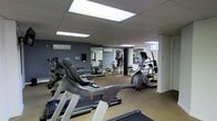 This fitness center offers a comprehensive workout environment designed to accommodate a range of exercise needs. The space is finished with neutral-toned flooring that provides a practical and durable surface throughout. Bold blue-grey accent walls add a modern, energizing atmosphere, while large mirrored panels along one wall visually expand the room and allow users to monitor their form during workouts. Overhead, recessed panel lighting ensures the entire area is brightly and evenly illuminated. The cardio section is well-appointed, featuring treadmills, an elliptical cross-trainer, and additional aerobic equipment arranged to allow comfortable spacing between machines. Toward the rear of the space, a dedicated strength training area includes a multi-station cable and weight machine, a workout bench, and free weights, offering users a full range of resistance training options. A wall-mounted split air conditioning unit helps maintain a comfortable temperature during intensive sessions. A small window near the strength area allows for a degree of natural light. The overall layout is efficient and functional, making this fitness center a practical and well-rounded facility for daily exercise routines.