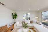 Located at 1512 Hollowhill Drive in Bryan, Texas, this single-floor living room offers a clean, modern aesthetic defined by its crisp white walls and warm hardwood flooring that runs throughout the space. The room is bathed in natural light, courtesy of a large, multi-panel picture window that frames a pleasant view of the surrounding neighborhood greenery, creating a seamless connection between the interior and the outdoors. Recessed ceiling lights complement the natural light, ensuring the space remains bright and welcoming at any time of day. The open layout provides a versatile setting, comfortably accommodating a plush sectional sofa paired with a sleek oval coffee table and a woven area rug that adds texture and warmth underfoot. A wicker accent chair and matching footstool introduce natural, organic elements, while a tall fiddle-leaf fig plant in the corner brings a refreshing touch of greenery indoors. Clean white baseboards and smooth wall finishes contribute to the room's polished, contemporary feel. The overall ambiance is calm, airy, and inviting — a well-designed living space that balances simplicity with comfort.