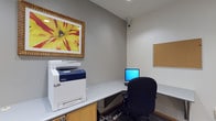 This single-floor business center is designed to provide a functional and efficient workspace environment. The main area features a continuous white laminate countertop that wraps along the walls, offering ample desk space for multiple users. A Xerox multifunction printer/copier is prominently stationed on one end of the counter, providing convenient print, scan, and copy capabilities. A desktop computer workstation with an ergonomic task chair sits nearby, accompanied by a wall-mounted corkboard for notices or reference materials. A security camera is mounted in the corner, ensuring the area is monitored. A vibrant framed floral artwork adorns the wall, adding a touch of color to the otherwise neutral gray palette.

Adjacent to the main area is a private workspace alcove, accessible through a wood-framed doorway. This quieter nook features an L-shaped white laminate desk, a task chair, and a laptop workstation, making it ideal for focused individual work. Built-in cabinetry with warm wood-toned finishes provides additional storage. The flooring throughout consists of patterned carpet tiles in muted gray tones, contributing to a professional atmosphere. Recessed lighting ensures consistent illumination across the space, creating a clean and productive environment overall.