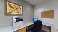 This single-floor business center is designed to provide a functional and efficient workspace environment. The main area features a continuous white laminate countertop that wraps along the walls, offering ample desk space for multiple users. A Xerox multifunction printer/copier is prominently stationed on one end of the counter, providing convenient print, scan, and copy capabilities. A desktop computer workstation with an ergonomic task chair sits nearby, accompanied by a wall-mounted corkboard for notices or reference materials. A security camera is mounted in the corner, ensuring the area is monitored. A vibrant framed floral artwork adorns the wall, adding a touch of color to the otherwise neutral gray palette.

Adjacent to the main area is a private workspace alcove, accessible through a wood-framed doorway. This quieter nook features an L-shaped white laminate desk, a task chair, and a laptop workstation, making it ideal for focused individual work. Built-in cabinetry with warm wood-toned finishes provides additional storage. The flooring throughout consists of patterned carpet tiles in muted gray tones, contributing to a professional atmosphere. Recessed lighting ensures consistent illumination across the space, creating a clean and productive environment overall.