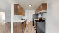 This single-floor unit offers a thoughtfully arranged layout that balances functionality with a clean, contemporary aesthetic. The galley-style kitchen is a standout feature, lined on both sides with rich, warm-toned wood cabinetry extending to the ceiling for ample storage. Light quartz countertops provide contrast, while stainless steel appliances — including a refrigerator, electric range with overhead exhaust hood, and dishwasher — complete the well-equipped cooking space. Light wood-look flooring runs throughout the kitchen and flows into the adjoining living areas, creating a cohesive feel. The living room is bright and open, featuring large windows dressed with vertical blinds that allow generous natural light, a wall-mounted air conditioning unit, and baseboard heating. An additional room with built-in open shelving and wood-look flooring offers flexible use as a dining area or home office. The bedrooms are carpeted for comfort, each featuring closets with bifold doors, baseboard heating, and windows that bring in natural light. The full bathroom is finished in a clean all-white palette, featuring a soaking tub with shower, wood-look flooring, a window with vertical blinds, and baseboard heating.