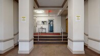 Located in the Dupont Circle neighborhood of Washington, DC, this building lobby at 1717 R St NW showcases classic early 20th-century architectural character. The entry vestibule greets visitors with a bold black-framed glass door flanked by symmetrical windows, allowing natural light to filter into the space and offering views of the tree-lined streetscape beyond. A cast-iron radiator positioned near the entrance reflects the building's period heating infrastructure, while ornate crown molding and decorative cornice details along the ceiling reinforce the traditional architectural aesthetic throughout.

A red runner carpet guides visitors from the front entry through the lobby, where the flooring transitions to a textured, neutral-toned surface. Structural columns frame the interior corridor, adding a sense of depth and formality to the layout. The lobby rises to a tiered landing area accessible via a short staircase fitted with metal handrails, accommodating accessibility needs on both sides. Warm wood-toned unit doors line the hallways, complementing the cream-painted walls. Overhead, flush-mounted light fixtures provide consistent illumination throughout. Fire safety equipment, including a visible sprinkler system and fire extinguisher, is thoughtfully integrated. The overall ambiance is functional yet dignified, preserving the architectural integrity of this historic Washington, DC address.