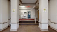 Located in the Dupont Circle neighborhood of Washington, DC, this building lobby at 1717 R St NW showcases classic early 20th-century architectural character. The entry vestibule greets visitors with a bold black-framed glass door flanked by symmetrical windows, allowing natural light to filter into the space and offering views of the tree-lined streetscape beyond. A cast-iron radiator positioned near the entrance reflects the building's period heating infrastructure, while ornate crown molding and decorative cornice details along the ceiling reinforce the traditional architectural aesthetic throughout.

A red runner carpet guides visitors from the front entry through the lobby, where the flooring transitions to a textured, neutral-toned surface. Structural columns frame the interior corridor, adding a sense of depth and formality to the layout. The lobby rises to a tiered landing area accessible via a short staircase fitted with metal handrails, accommodating accessibility needs on both sides. Warm wood-toned unit doors line the hallways, complementing the cream-painted walls. Overhead, flush-mounted light fixtures provide consistent illumination throughout. Fire safety equipment, including a visible sprinkler system and fire extinguisher, is thoughtfully integrated. The overall ambiance is functional yet dignified, preserving the architectural integrity of this historic Washington, DC address.