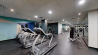 Located at 5901 Montrose Rd in Rockville, MD, this well-appointed fitness center spans a generous single-floor layout designed to accommodate a full range of workout activities. The space is organized into distinct functional zones, each thoughtfully equipped to serve different training needs.

The cardio area features a row of treadmills and elliptical machines, positioned beneath a drop-panel ceiling with recessed lighting that maintains a bright, energizing atmosphere. Adjacent to this section, a dedicated strength training zone offers an extensive array of selectorized machines, free weights, barbells, and benches, complemented by floor-to-ceiling mirrors that enhance the sense of space and allow for proper form monitoring.

A separate cycling studio provides a focused environment for indoor cycling classes, featuring multiple stationary bikes with integrated screens, a hardwood floor, deep teal-painted walls, and a full-length mirror. Glass partition doors connect this room to the main floor, maintaining visual openness while offering acoustic separation.

Throughout the facility, bold geometric accent murals in navy, teal, and white add a modern, motivating aesthetic. Large windows along the perimeter bring in natural light and views of the surrounding greenery. Dark rubberized flooring in the main areas ensures durability and safety, while the overall layout provides ample circulation space between equipment stations.