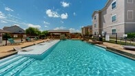 Located at 1101 Longhorn Rd in Fort Worth, Texas, this outdoor swimming pool area is a well-appointed amenity space designed for relaxation and recreation under the Texas sun. The centerpiece is a generously sized rectangular pool with crystal-clear blue water, complemented by elegant stone water features that cascade gently into the pool, adding both visual appeal and a soothing ambiance.

The expansive pool deck is finished in a warm tan concrete surface, providing ample space for lounging. Rows of teal-cushioned chaise lounge chairs are arranged along the perimeter, offering plenty of room for sunbathing. A raised spa or wading area is integrated into the pool design, further enhancing the resort-like experience.

A covered pavilion with a pitched metal roof and warm wood ceiling anchors one end of the deck, providing shaded seating and an outdoor kitchen or grill area constructed from matching limestone-style stonework. The enclosure is secured by black wrought-iron fencing with stone pillar accents, flanked by decorative barrel planters filled with tropical greenery and colorful flowering plants. The surrounding residential buildings feature a neutral stucco and stone exterior, framing the pool area with a cohesive architectural aesthetic. Open views of the sky and distant treeline complete the inviting outdoor atmosphere.