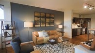 This single-floor space showcases a well-appointed living area that blends contemporary design with warm, inviting elements. The living room is anchored by rich hardwood flooring and a striking dark slate-blue accent wall, which serves as a dramatic backdrop for a large grid-style artwork arrangement — a standout feature that adds visual depth and character to the room. A tan leather sofa, dressed with decorative pillows and a soft throw blanket, sits atop a geometric patterned area rug, complementing the warm tones of the flooring. A dark rectangular coffee table anchors the seating arrangement, while two floor and table lamps provide ambient lighting throughout the space. The open layout connects the living area seamlessly to a kitchen visible in the background, which features dark cabinetry and modern appliances, contributing to the cohesive, contemporary aesthetic. White trim and neutral-toned ceilings contrast neatly with the bold accent wall, creating a balanced and polished interior. The overall ambiance is modern yet comfortable, with a carefully curated palette of warm browns, deep grays, and crisp whites that give the space a sophisticated and livable feel.