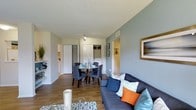 This single-floor space offers a thoughtfully designed open-plan layout that seamlessly connects the living and dining areas, creating a functional and inviting environment. The living area is anchored by a plush sofa adorned with coordinating throw pillows in navy, teal, and orange tones, complemented by a glass-top coffee table that adds a contemporary touch. Warm wood-style flooring runs throughout, providing a cohesive and welcoming aesthetic that pairs well with the soft grey-blue wall tones. The dining area features a round table surrounded by upholstered chairs, making it an ideal setting for both casual meals and entertaining. A track light fixture above the dining space ensures focused, practical illumination. The textured ceiling adds depth and character to the interior. Towards the rear of the space, multiple closeted storage areas with louvered and sliding doors offer generous organizational capacity. A hallway leads to additional rooms, providing natural separation between living and private areas. Framed wall art and decorative accents contribute to the polished, curated feel of the space, while a glimpse of an adjoining kitchen hints at further functional amenities. Overall, the space balances comfort, style, and practicality in a well-organized single-level layout.