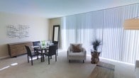 This single-floor space combines a living and dining area in an open-plan layout that feels spacious and well-lit. The most prominent architectural feature is the expansive floor-to-ceiling window dressed with white vertical blinds, which diffuse natural light throughout the room and create a soft, luminous ambiance.

The dining area, positioned to one side, accommodates a dark-toned glass-top table paired with matching black chairs, offering a sleek and contemporary dining setup. A full-length mirror in a dark frame leans against the wall nearby, adding depth and visual interest to the corner.

The living area features a light-grey upholstered armchair accented with a decorative patterned cushion, complemented by a distinctive side table with a circular glass top supported by a natural driftwood base — a standout organic element that adds texture and character to the space.

Neutral beige carpeting covers the main floor area, contributing to the warm and understated tone of the interior. The textured ceiling and soft wall tones further enhance the clean, contemporary feel of the space overall.