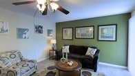 This single-floor living space centers around a well-appointed living room that combines comfort with thoughtful design. A striking olive green accent wall serves as a bold focal point, contrasting with the surrounding light-toned walls to create visual depth and warmth. Three framed abstract artworks are displayed prominently against the green wall, adding a curated, gallery-like quality to the space.

The room is furnished with a dark upholstered sofa and a coordinating accent chair, both accented with decorative patterned pillows. A round wooden coffee table anchors the seating area atop a contemporary area rug featuring a circular motif in neutral tones. A small side table with a table lamp provides ambient lighting alongside the ceiling fan with a built-in light fixture, which serves as the room's central overhead lighting source.

The textured ceiling adds subtle architectural character, while the light-colored carpeted flooring ties the elements of the room together. Decorative accessories, including teal candle holders and colorful orbs on the coffee table, introduce playful accents throughout the space, contributing to an overall ambiance that feels both inviting and stylishly composed.
