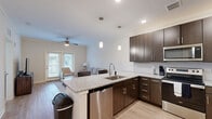 This well-appointed one-bedroom, one-bathroom apartment at Cool Springs Drive in Elizabethtown, Kentucky offers a thoughtfully designed single-floor layout. The open-concept living and kitchen area serves as the heart of the home, where rich dark-wood cabinetry, granite countertops, and a full suite of stainless steel appliances—including a refrigerator, range, microwave, and dishwasher—create a modern, functional kitchen. A peninsula with seating provides a casual dining option, accented by stylish pendant lighting overhead. The living room features crown molding, a ceiling fan, and light hardwood-style flooring, with a glass door opening to outdoor access and allowing natural light to fill the space. The bedroom is a comfortable retreat with plush carpeting, a ceiling fan, and a large window framing outdoor views. Multiple closets, including a dedicated laundry space and hallway storage, offer generous organization options throughout. The bathroom is clean and neutral, featuring a granite-topped vanity with dark cabinetry, a full tub-shower combination, and wood-style flooring. Recessed lighting and a consistent neutral color palette throughout give the entire apartment a bright, cohesive feel.