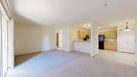 This single-floor unit is designed with a practical, open-concept layout that connects the living area directly to the kitchen. The living space is generously sized, finished with neutral-toned wall-to-wall carpeting and cream-colored walls that lend a clean, bright atmosphere throughout. A peninsula counter separates the kitchen from the main living area, offering a functional transition between spaces. The kitchen is equipped with light wood cabinetry, a black refrigerator, and a microwave, all set beneath flush-mount ceiling lighting.

Multiple additional rooms branch off from the main living area, each carpeted and featuring baseboard heating units for consistent climate control. One room benefits from a large multi-pane window fitted with horizontal blinds, allowing ample natural light to filter in while providing views of surrounding greenery. Another room includes a wall-mounted utility panel and baseboard heating, offering versatility in use.

The bathroom is finished with large-format tile flooring and includes a full soaking tub with grab bars, a light wood vanity with storage, and a towel bar. The overall palette of the unit is consistently neutral, with cream walls, light trim, and carpeted floors creating a cohesive and understated aesthetic throughout.