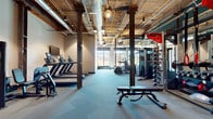 Located at 96 E Catawba St in Belmont, NC, this fitness facility blends industrial character with modern functionality. The space features an exposed wood-plank ceiling with visible ductwork and structural columns, lending it a distinctive loft-like aesthetic that sets it apart from conventional gyms.

The main floor is thoughtfully organized into dedicated workout zones. A cardio section lines one wall, housing a row of treadmills equipped with individual monitors and a wall-mounted flat-screen display. An elliptical trainer and additional cardio equipment are positioned near large windows that frame outdoor views and flood the space with natural light.

The strength training area is well-stocked with a multi-station cable machine, a full dumbbell rack, kettlebells, medicine balls, stability balls, and a power rack system with attachments — offering a comprehensive range of resistance training options. An adjustable weight bench provides a versatile focal point in the center of the floor.

A separate studio room, accessible through striking teal-framed glass double doors, offers a clean, open environment suitable for stretching, yoga, or personal training sessions. A beverage cooler is conveniently positioned near the studio entrance. The flooring throughout consists of durable carpet tile, and ceiling fans contribute to airflow across the training floor.